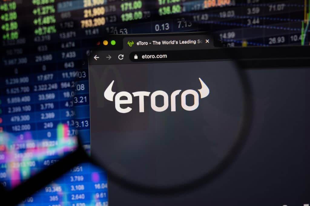 eToro company logo on a website with blurry stock market developments in the background, seen on a computer screen through a magnifying glass